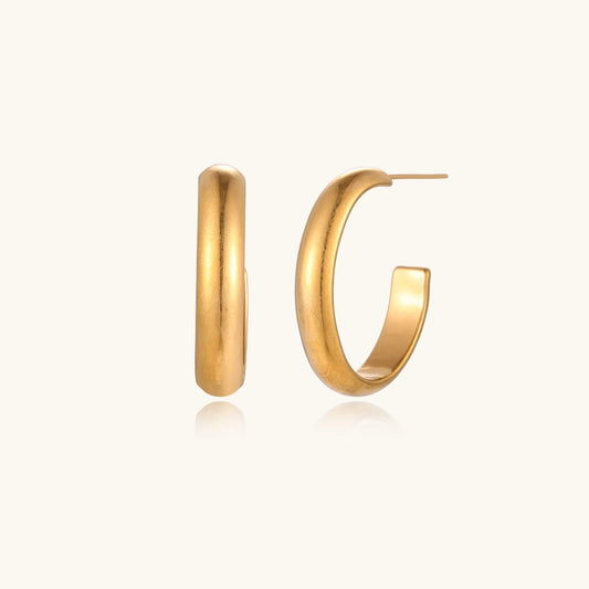 Chunky tubular hoop earrings in warm 14K gold-plated finish with smooth polished surface on cream background.