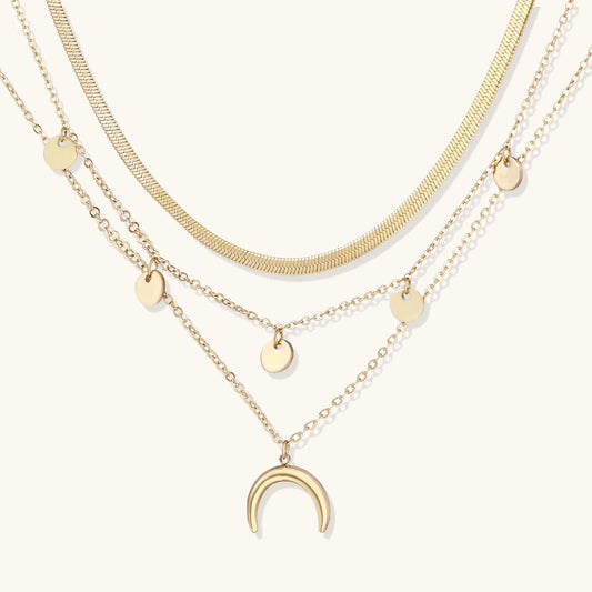 Four-layer gold necklace with herringbone chain, disc charms, and crescent moon pendant on cream background