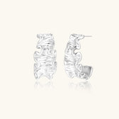 White gold plated sculptural hoop earrings with organic wavy edges and reflective metallic finish on cream background