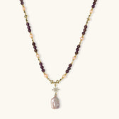 Gold necklace with iridescent baroque pearl pendant, purple amethyst beads, and crystal star charm on cream background